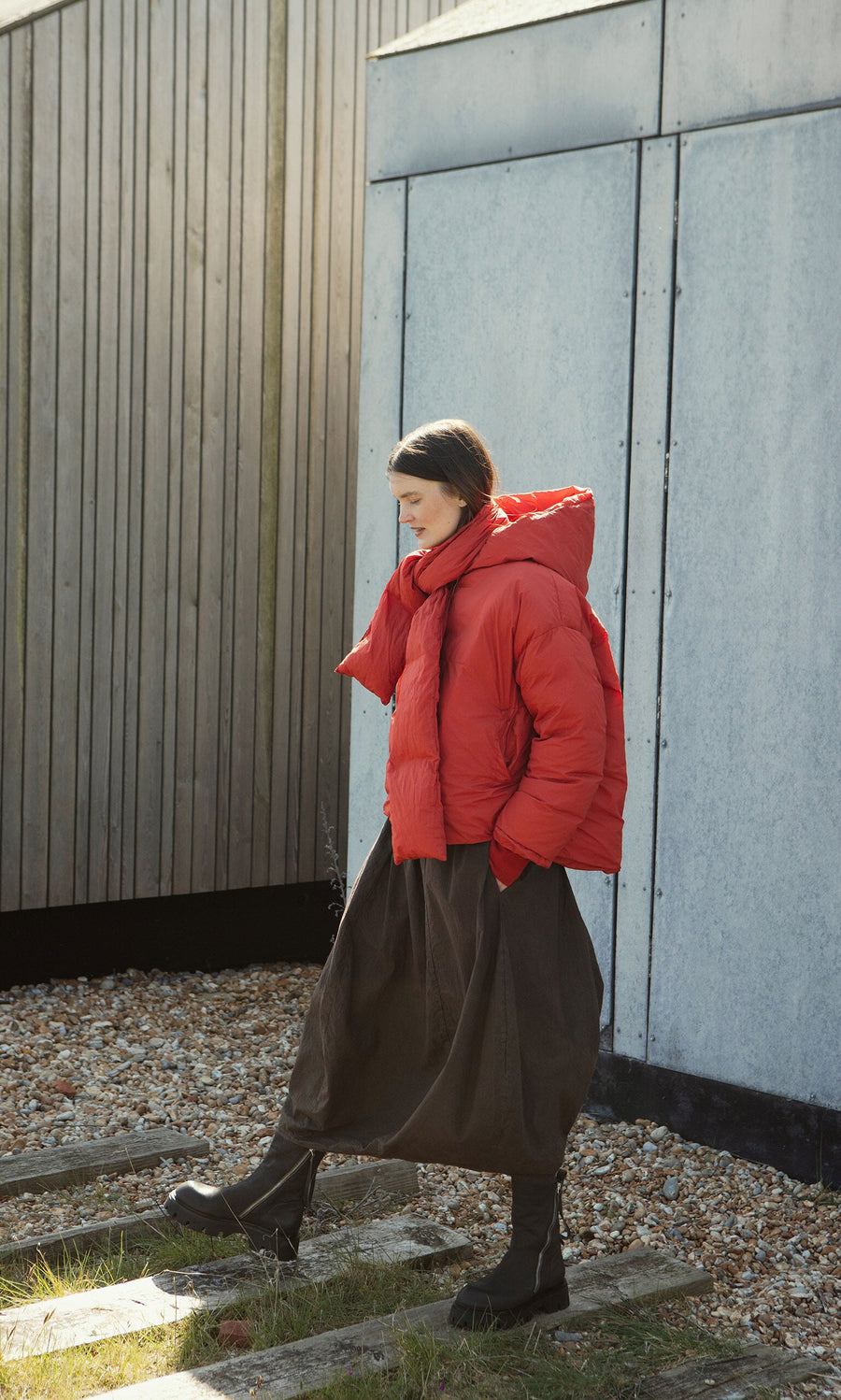 Plümo- Woman in a Red Puffer coat stands outside by a wooden and metal building, wearing a long dark skirt.