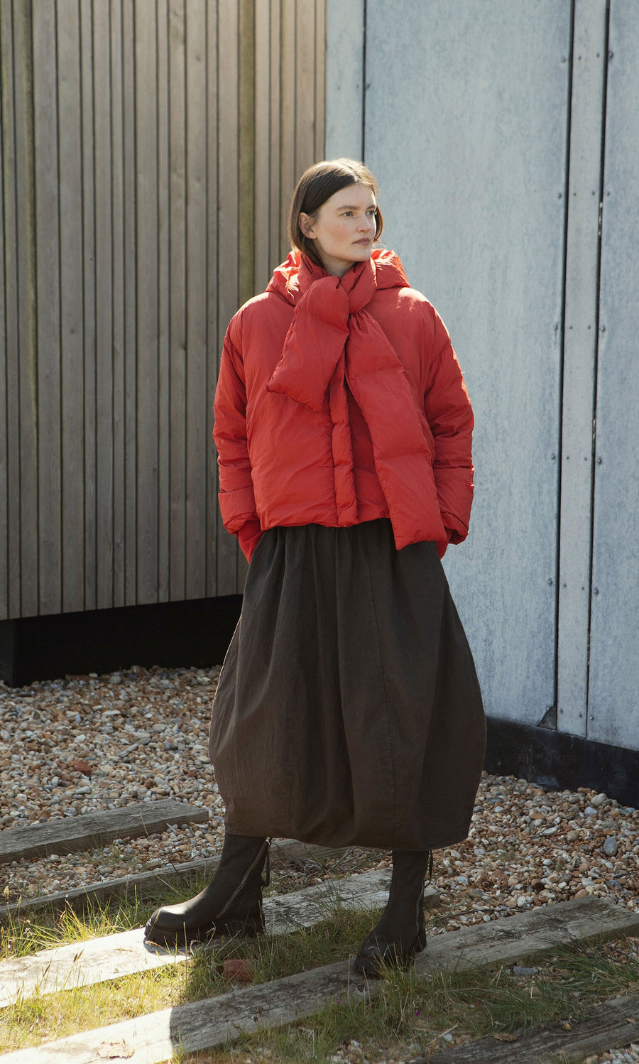 Plümo- A person in a red puffer coat, dark skirt, and boots stands by wooden and concrete walls.