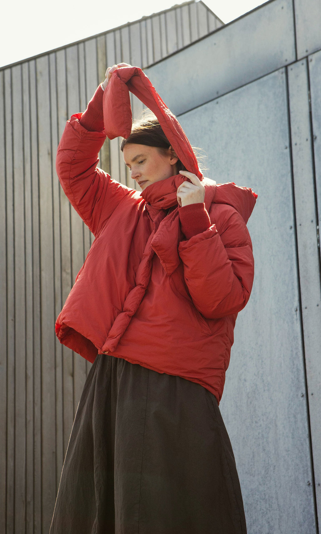 Plümo- Wearing a Red Puffer coat, a person lifts the hood over their head, standing by a modern metal and wood wall.