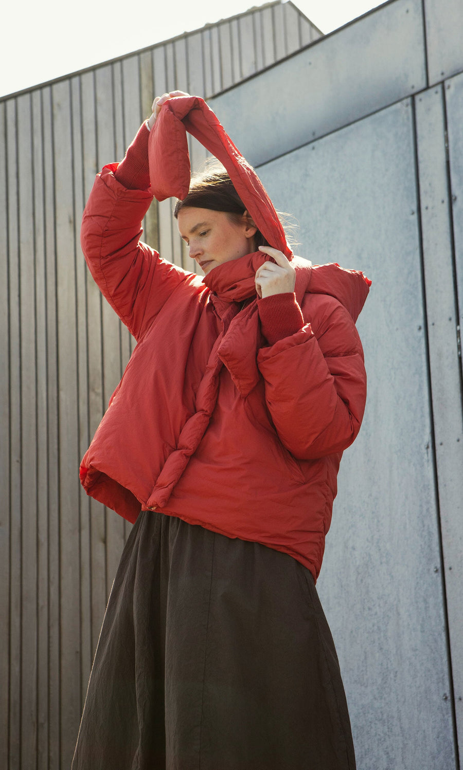 Plümo- Wearing a Red Puffer coat, a person lifts the hood over their head, standing by a modern metal and wood wall.