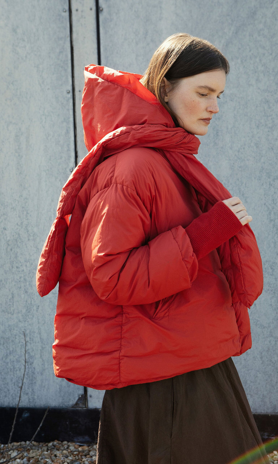 Plümo- Woman stands sideways in a red Puffer coat with detachable scarf and brown skirt, eyes closed against gray metal.