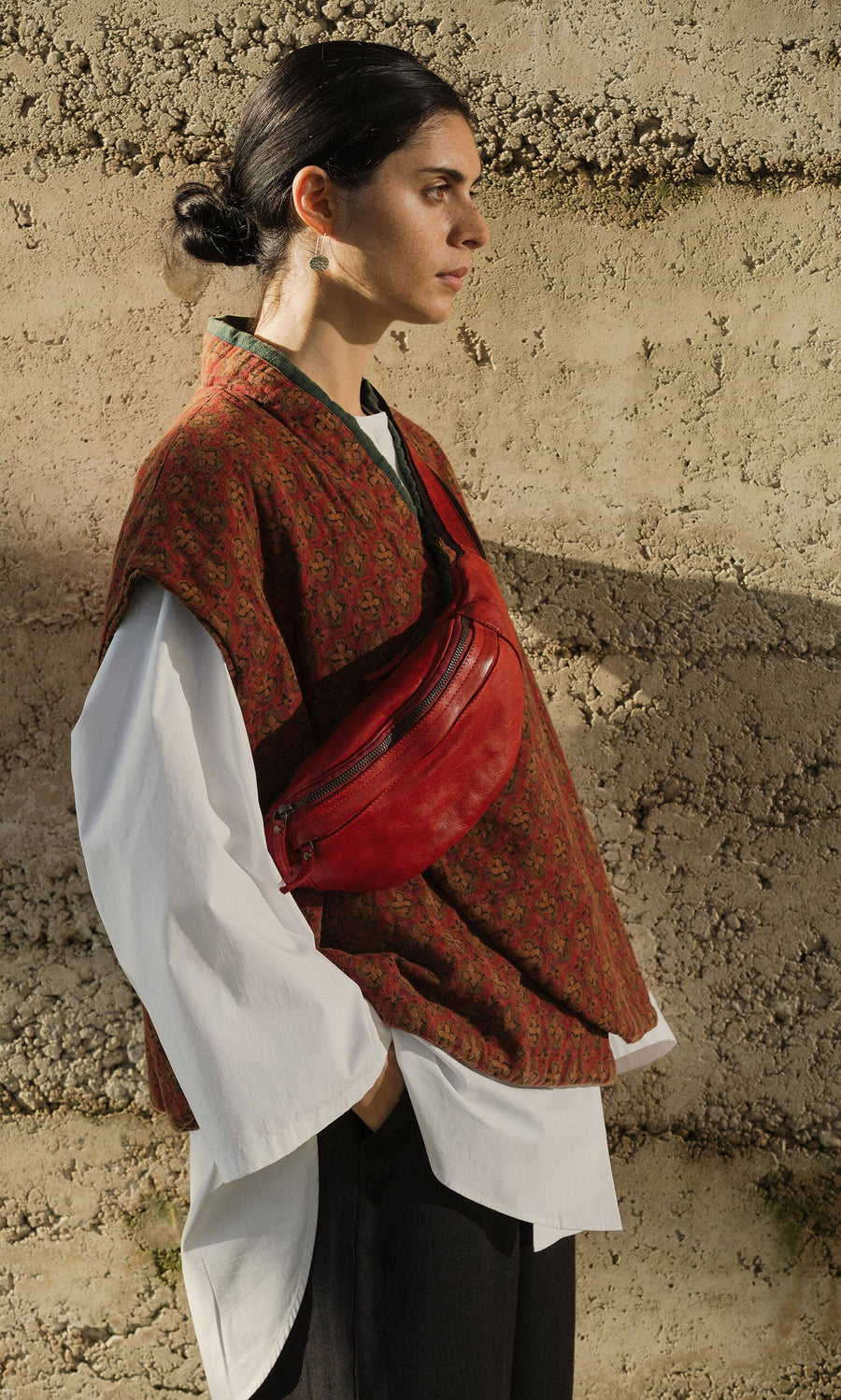 Plümo- Woman with dark hair in a bun, layered outfit, wears the Redwick Bag bumbag against a textured wall.