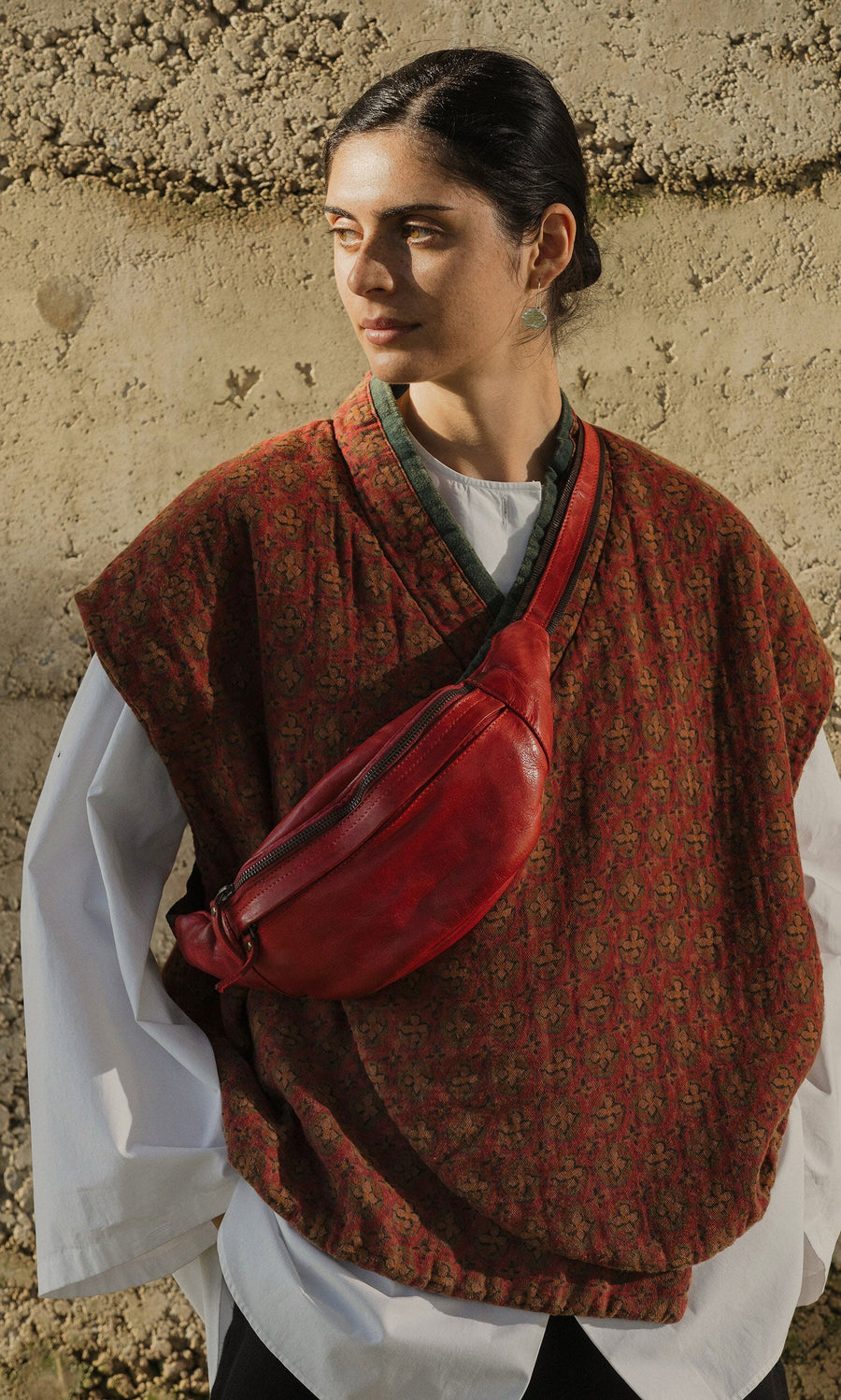 Plümo- Woman in patterned vest and white shirt stands against a textured wall, wearing the Redwick Bag crossbody.