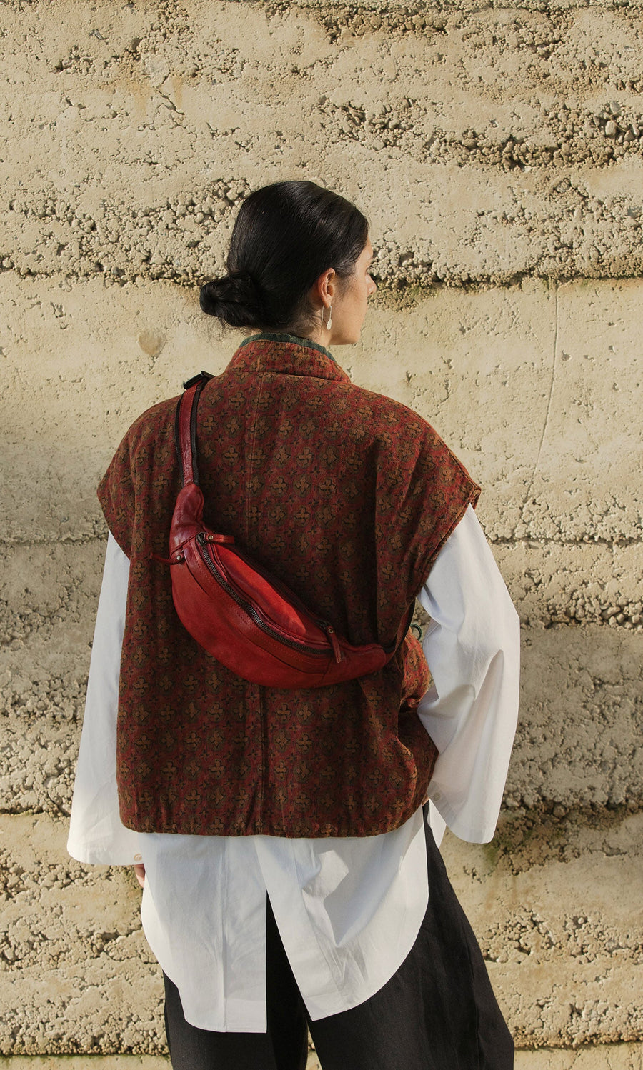 Plümo- Person with dark hair in a bun wears a British design vest and the Redwick Bag, standing by a textured wall.