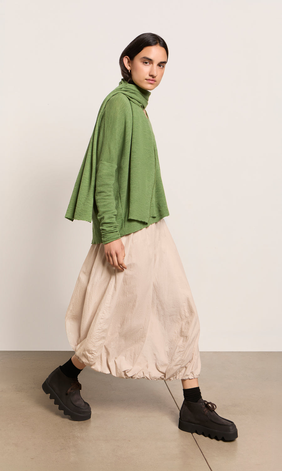 Plümo  Person in green top, beige balloon skirt, Renee Scarf, and black platforms walking indoors against a plain background.