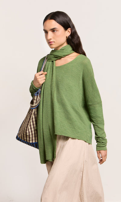 Plümo  Person in a green sweater with the Renee Scarf, beige skirt, and checkered bag, walking against a plain background.