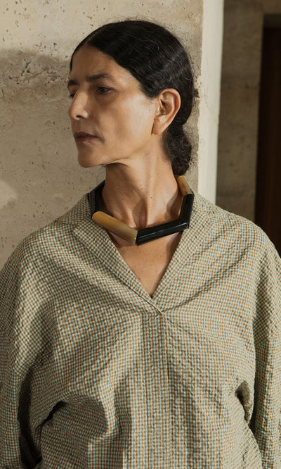 Plümo- Woman with dark hair in a low bun, wearing a checked shirt and the Rhia Necklace, stands by a stone wall.