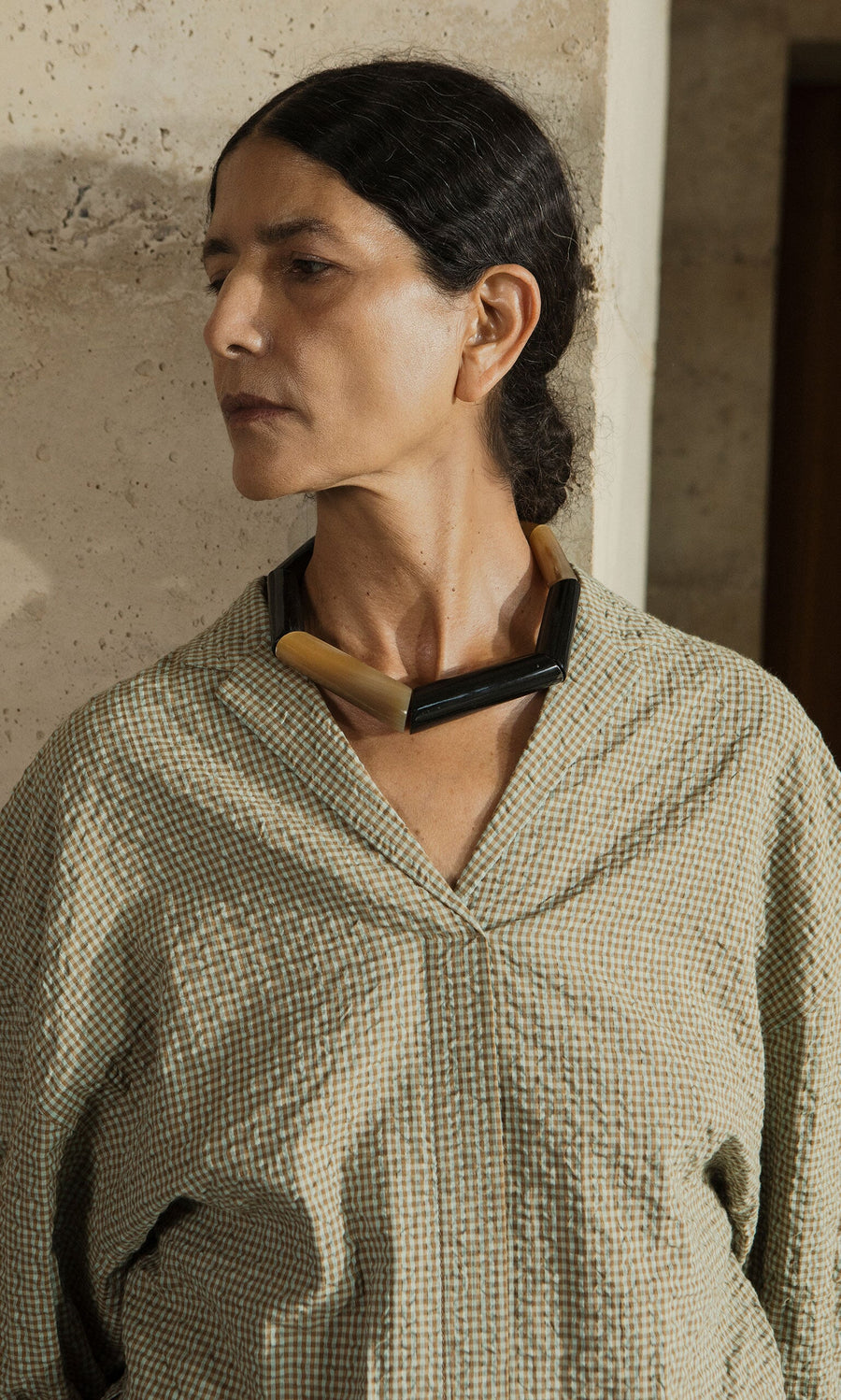 Plümo- Woman with dark hair in a low bun, wearing a checked shirt and the Rhia Necklace, stands by a stone wall.