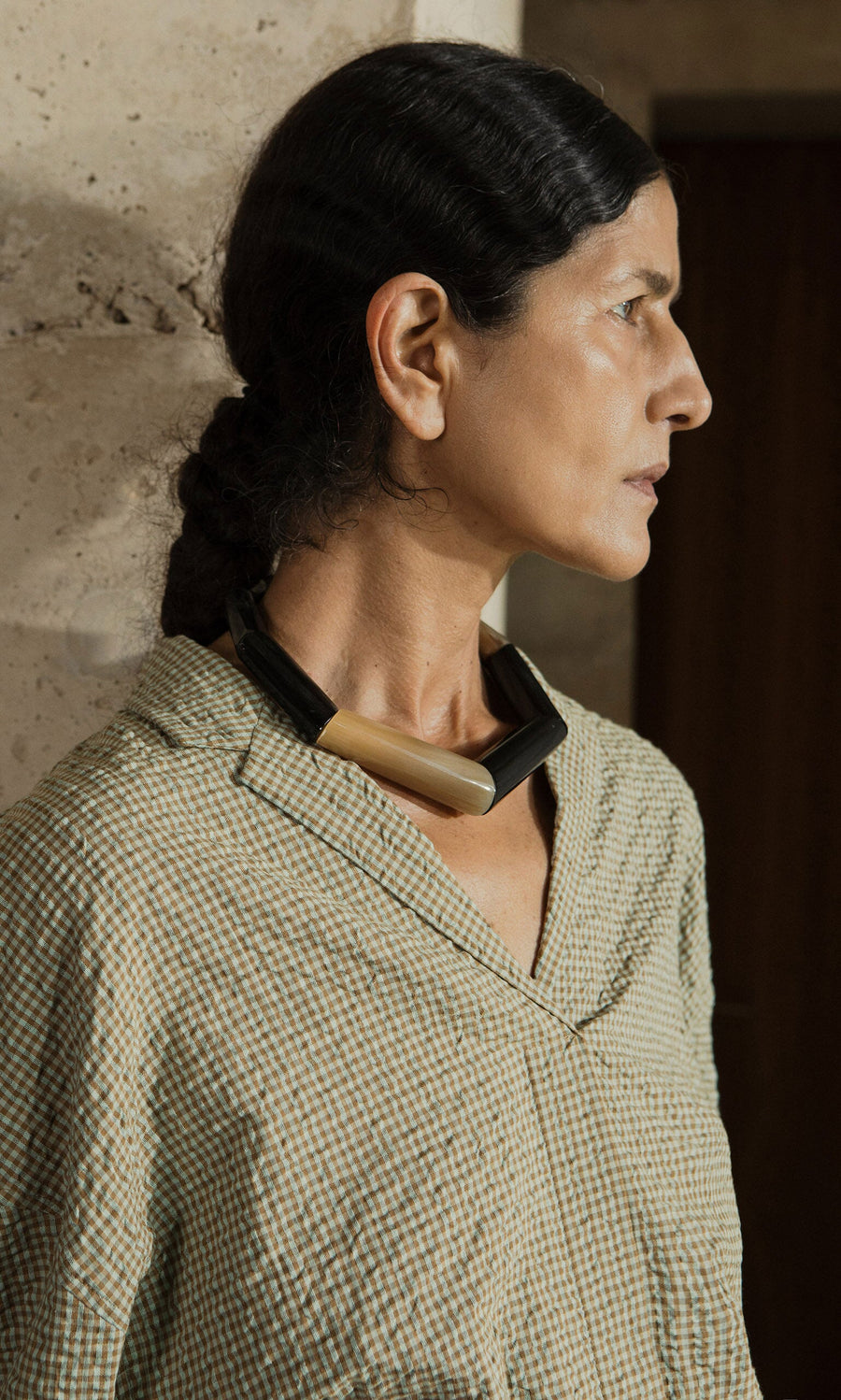 Plümo- Woman with dark hair in a low bun, wearing a green checked shirt and the Rhia Necklace, looks to the side.
