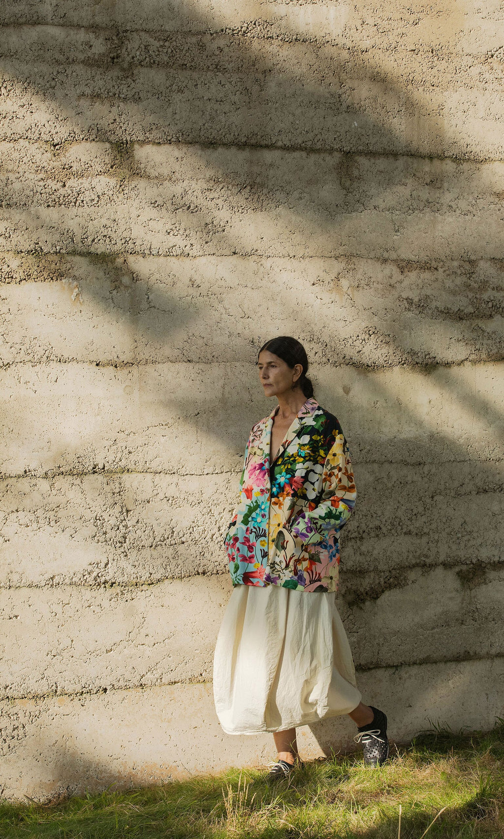 Plümo- Person in a hand-painted Riverene Blazer and white skirt stands by a textured concrete wall as sunlight casts shadows.