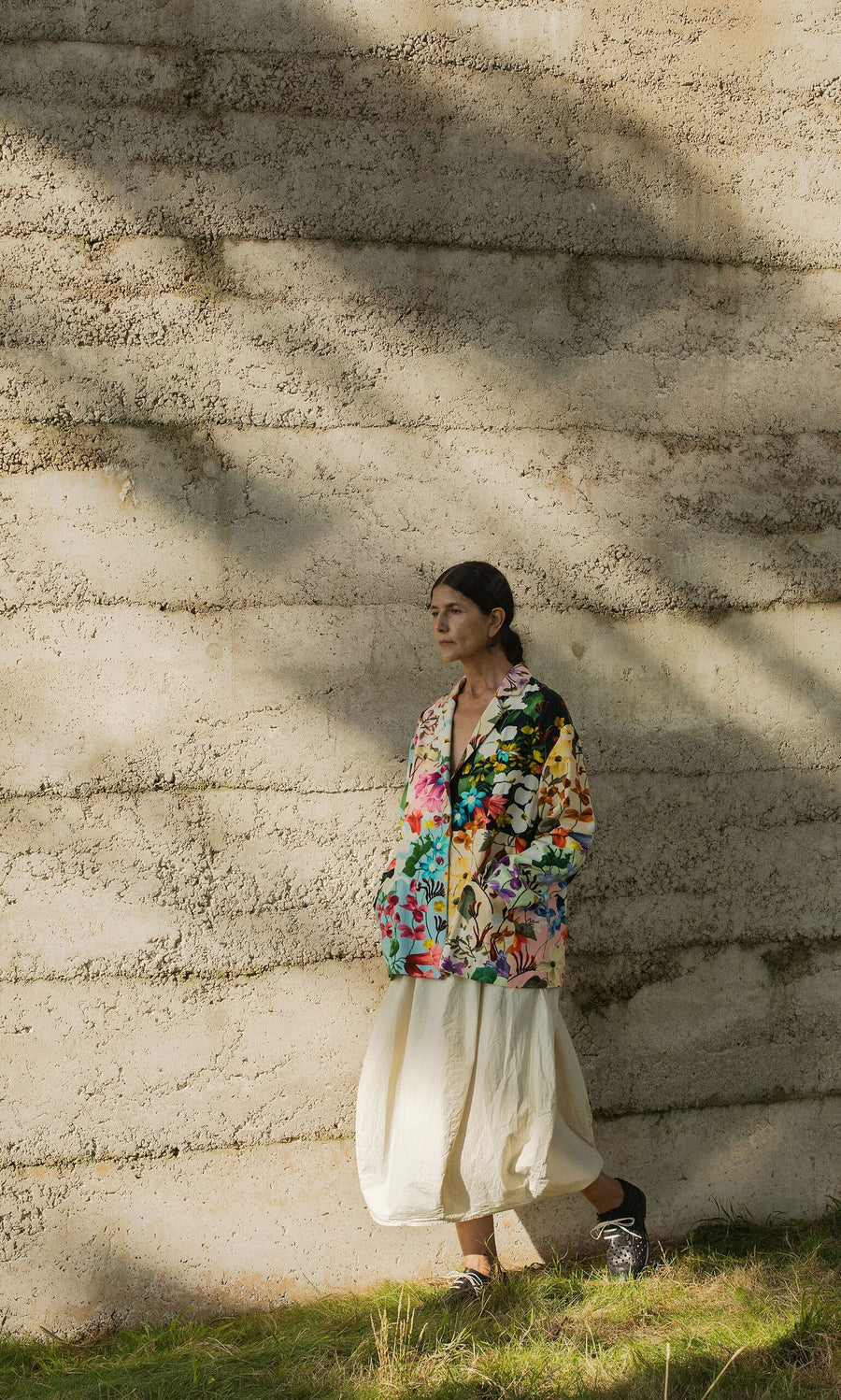 Plümo- Person in a hand-painted Riverene Blazer and white skirt stands by a textured concrete wall as sunlight casts shadows.