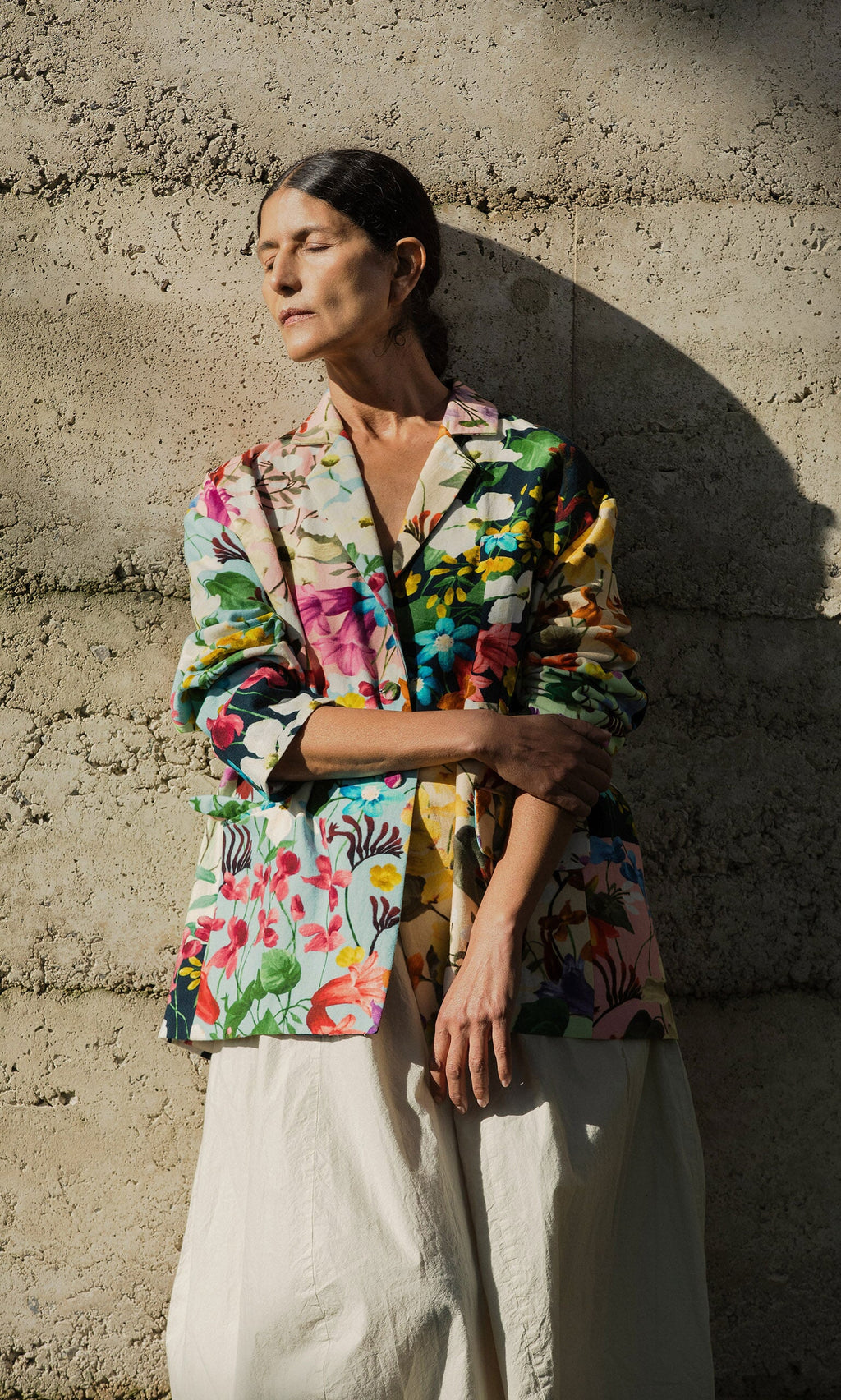 Plümo- Woman in a Riverene Blazer and cream skirt stands against a textured wall, eyes closed in sunlight and shadow.