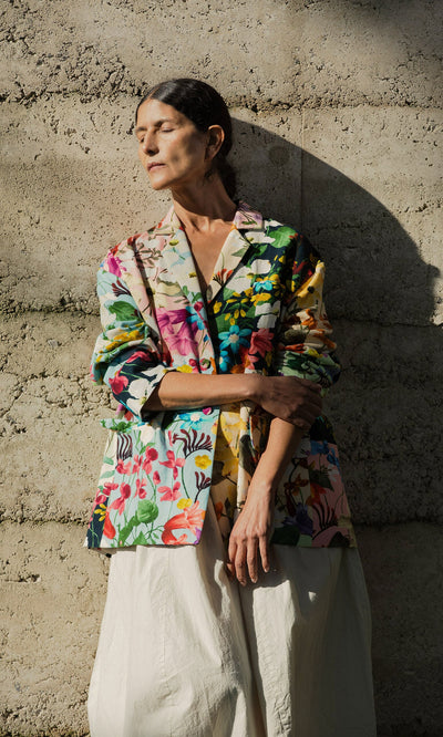 Plümo- Woman in a Riverene Blazer and cream skirt stands against a textured wall, eyes closed in sunlight and shadow.