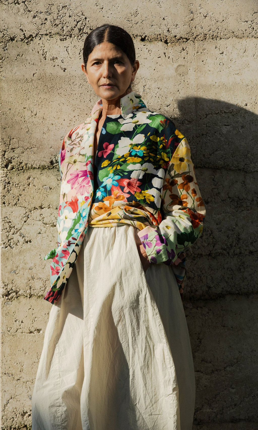 Plümo- Wearing the Riverene Blazer and cream skirt, a person stands against a textured concrete wall in sunlight.