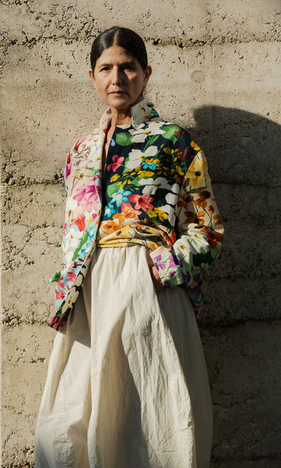 Plümo- Wearing the Riverene Blazer and cream skirt, a person stands against a textured concrete wall in sunlight.