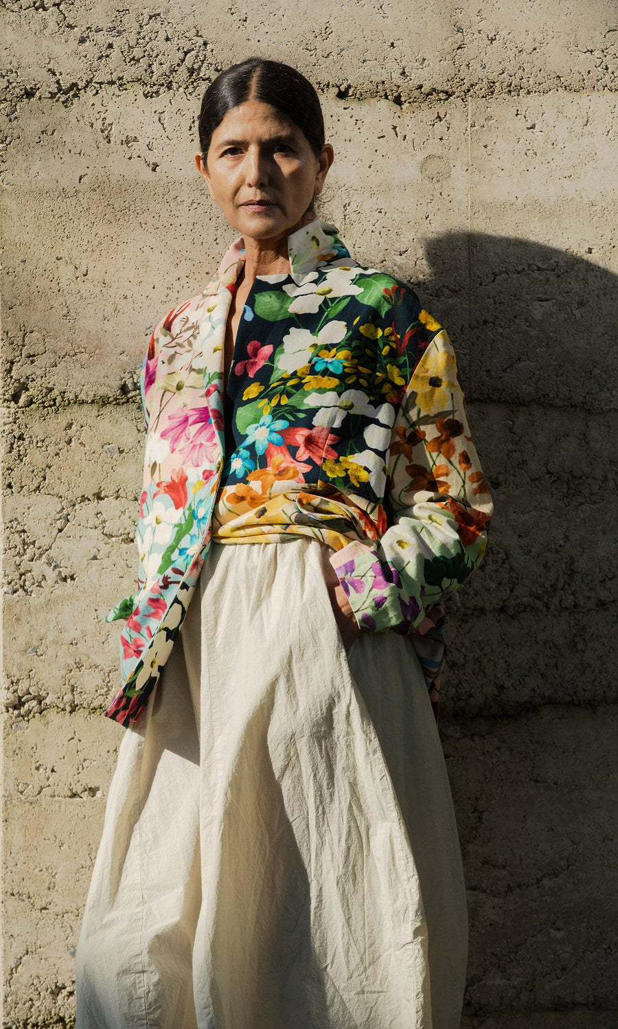 Plümo- Wearing the Riverene Blazer and cream skirt, a person stands against a textured concrete wall in sunlight.