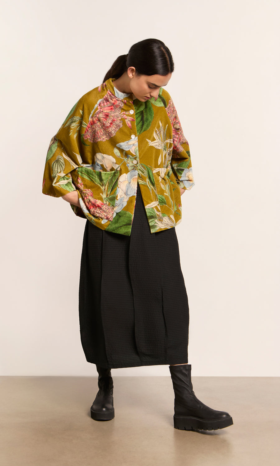 Plümo  Woman in a Rodart Velvet Jacket, black skirt, and boots stands looking down with hands in her pockets.