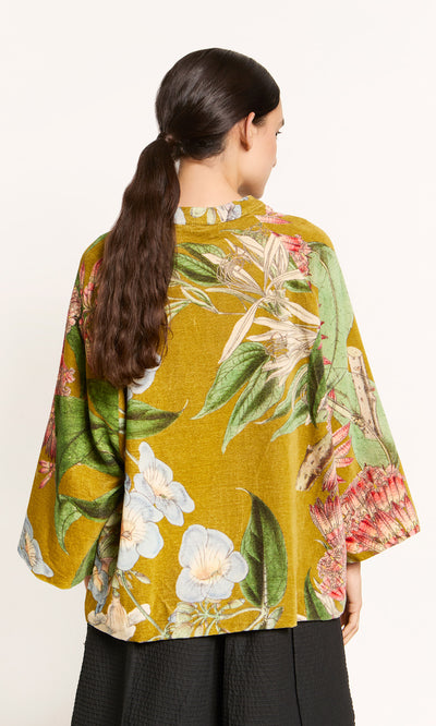Plümo  Woman with long brown ponytail wears Rodart Velvet Jacket in yellow botanical print, shown from the back.