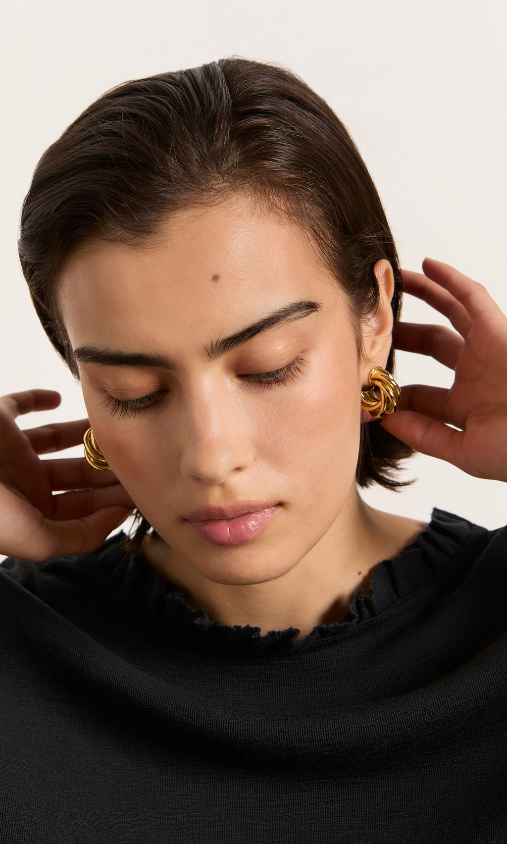 Plümo  Woman with short dark hair and eyes closed adjusts her hair, wearing Banda Earrings and a black top.