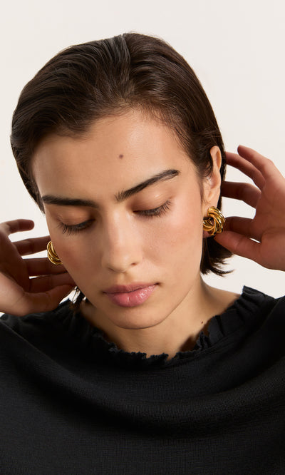 Plümo  Woman with short dark hair and eyes closed adjusts her hair, wearing Banda Earrings and a black top.