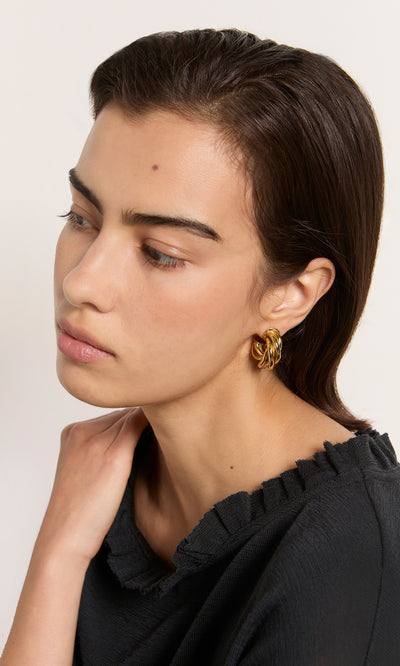 Plümo  Woman with dark hair wears Banda Earrings and a black top, looking down with a neutral expression.