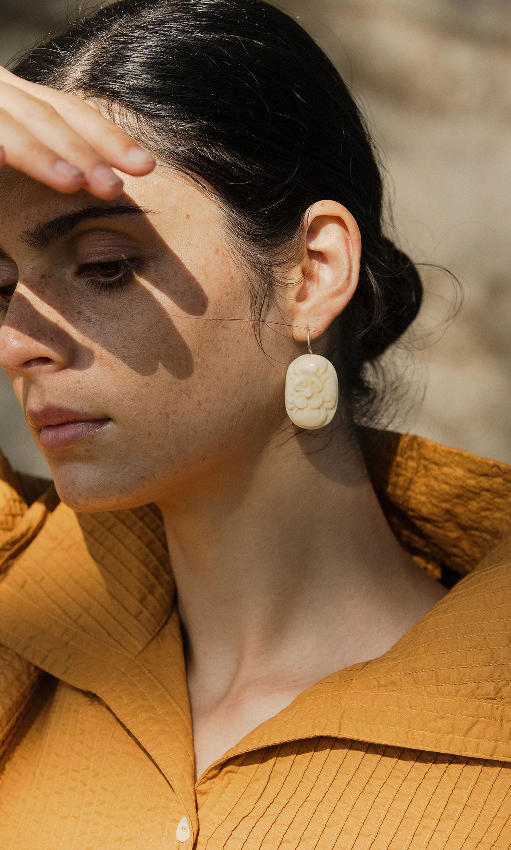Plümo- A woman in a textured mustard shirt shades her face, wearing the delicate floral motif Rose Earrings.