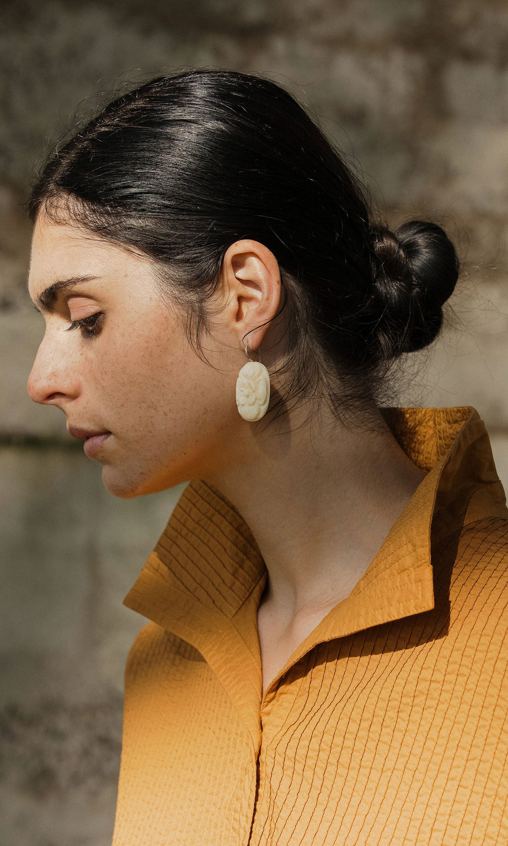 Plümo- Woman with dark hair in a low bun, wearing a textured mustard shirt and Rose Earrings, looking down.