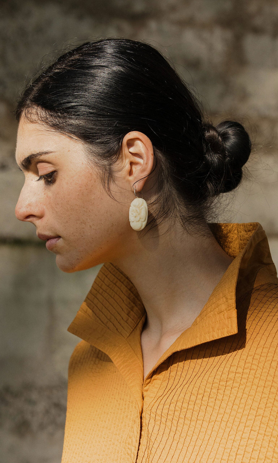 Plümo- Woman with dark hair in a low bun, wearing a textured mustard shirt and Rose Earrings, looking down.