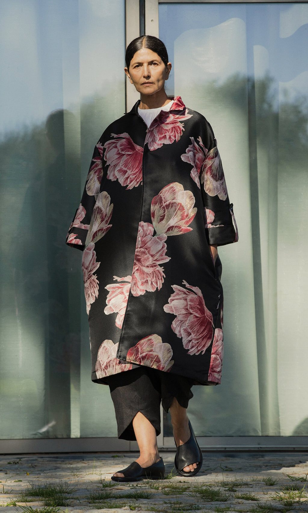 Plümo- Woman wearing the Rozella Coat, black pants, and shoes while standing outdoors by glass doors.