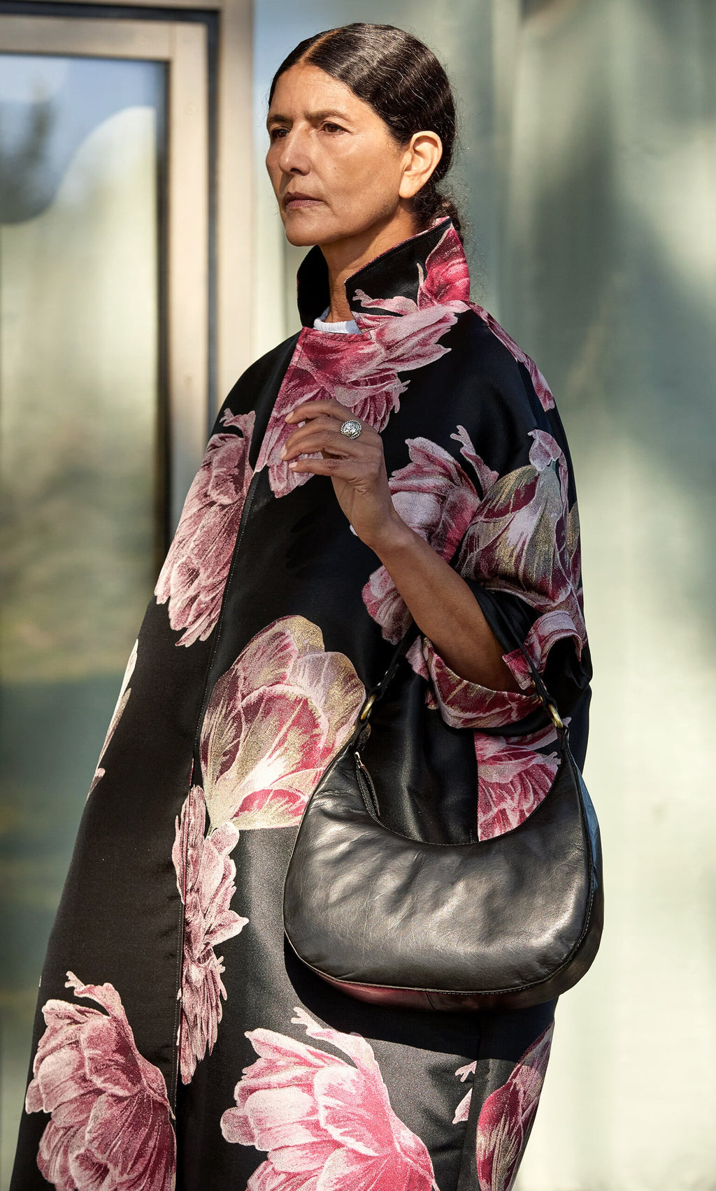 Plümo- Woman in a black floral coat holds the Hadlow Bag outdoors, sunlight illuminating her face.