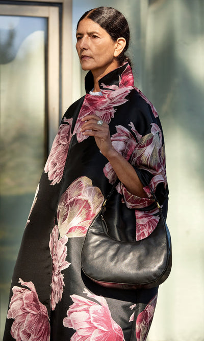 Plümo- Woman in a black floral coat holds the Hadlow Bag outdoors, sunlight illuminating her face.