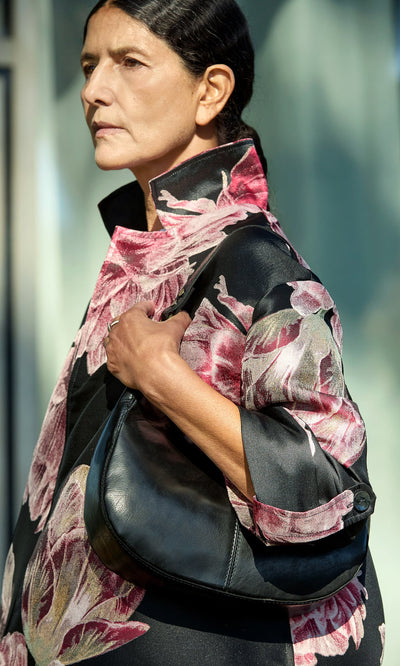 Plümo- Woman outdoors in a floral coat with a high collar, carrying the Hadlow Bag—crescent-shaped polished leather—over her shoulder.