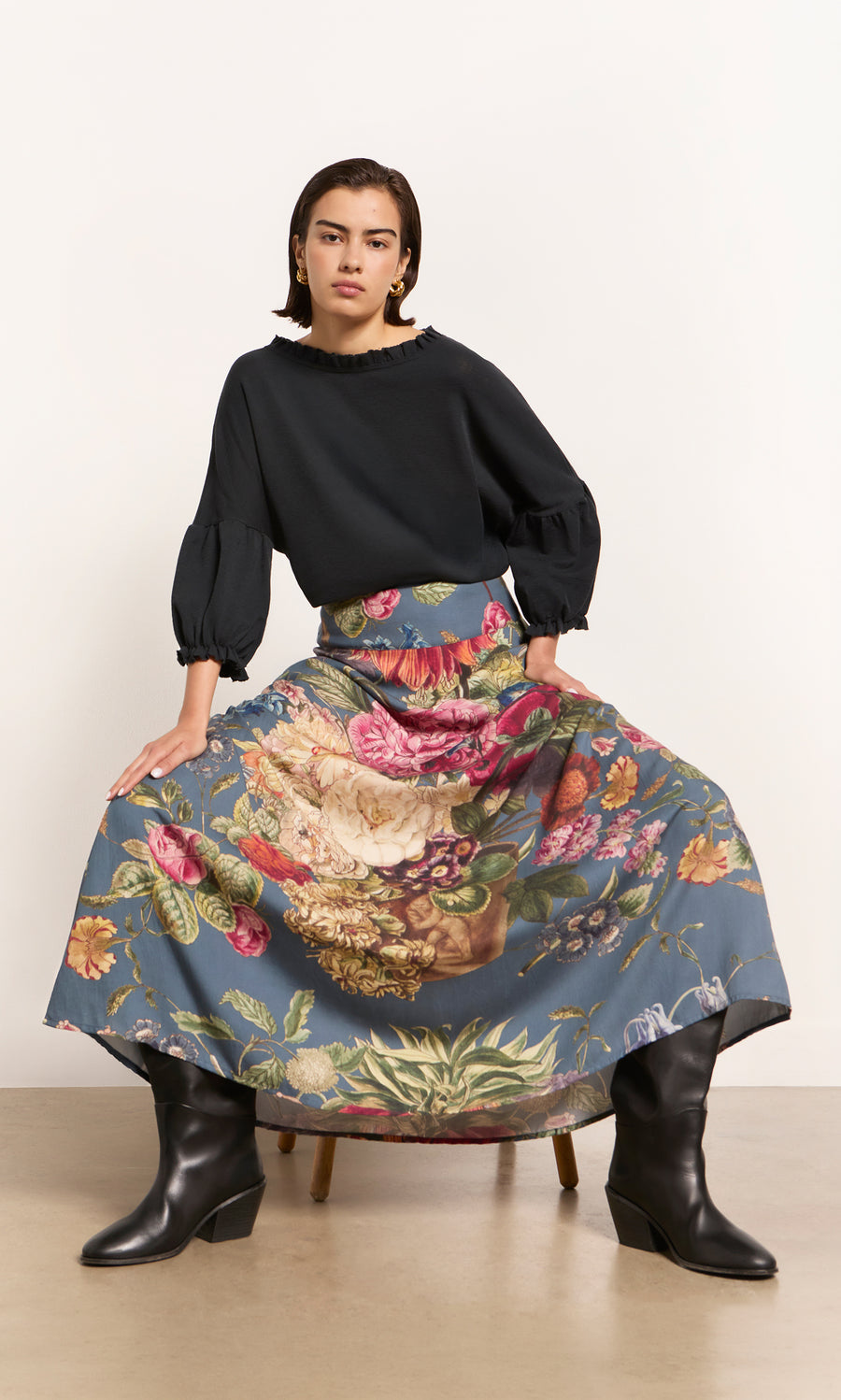 Plümo  Person in the Ruffle Top – Caviar, floral skirt, and boots sits on a chair against a plain background.