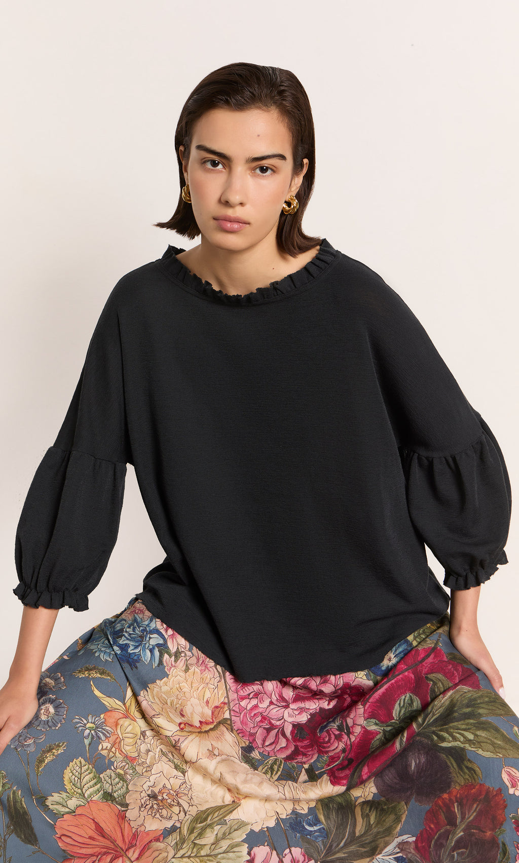 Plümo  Woman in a black ruffled top, floral skirt, and Banda Earrings sits against a plain background, facing the camera.