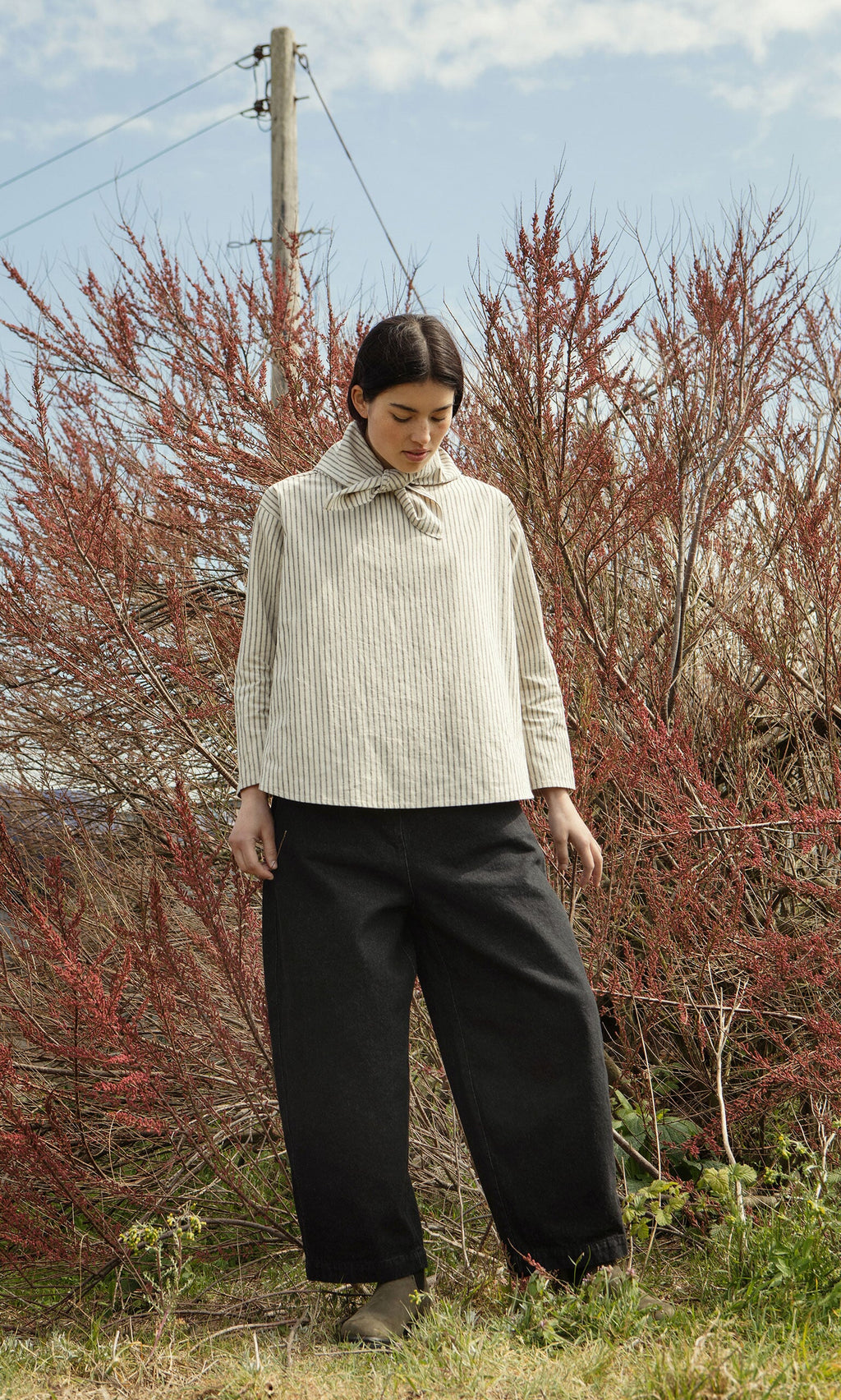 Plümo- A person in a Sail Cloth Top and wide black pants stands outdoors before tall reddish bushes under a cloudy sky.