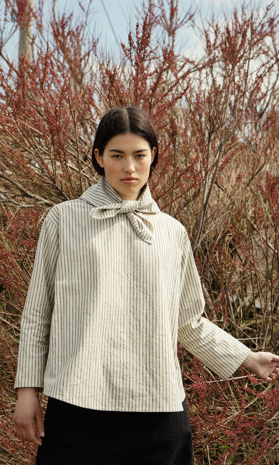 Plümo- Woman in the Sail Cloth Top with bow collar stands outdoors before red leafless bushes. Made in London.