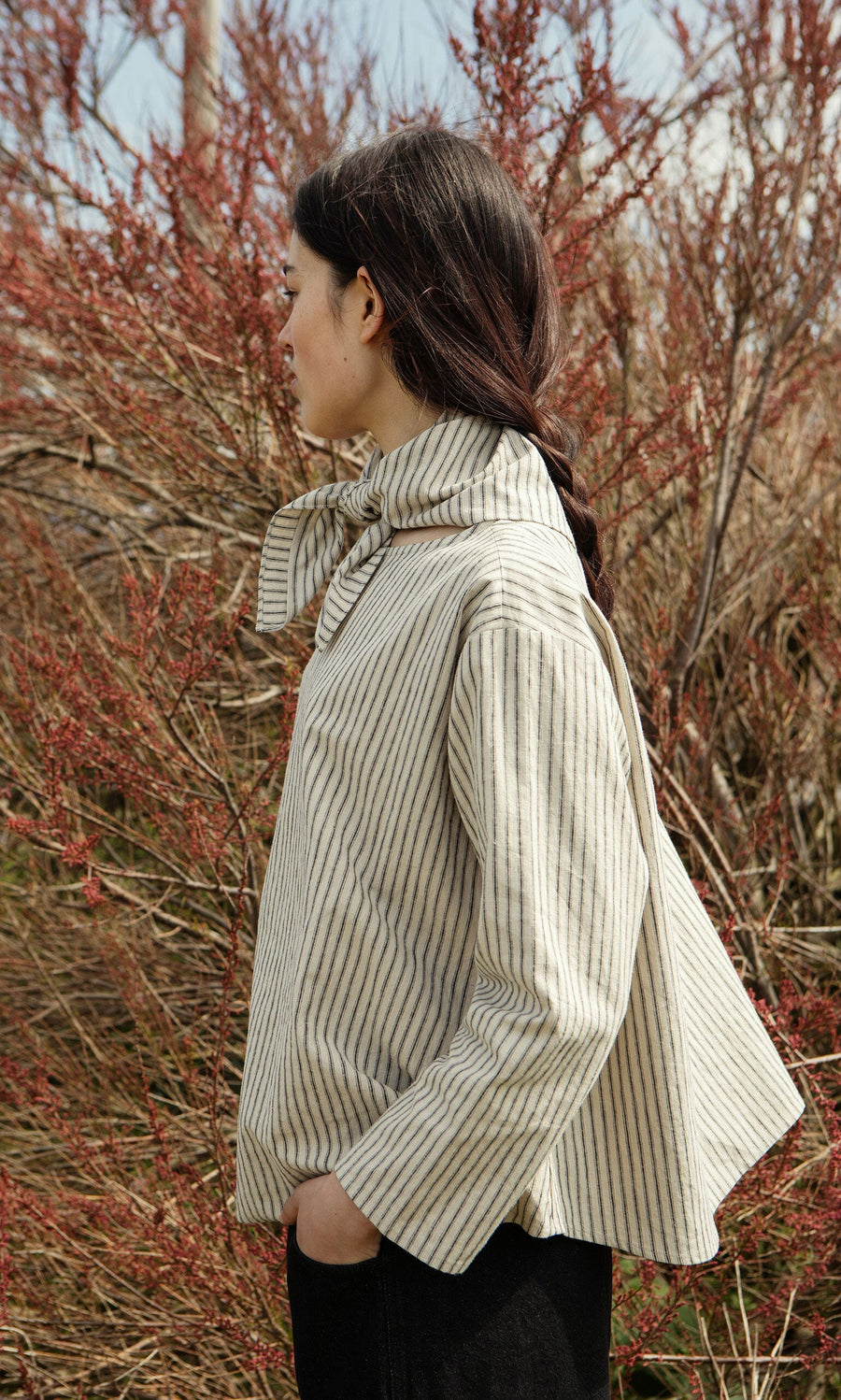 Plümo- Woman with dark braided hair wears a Sail Cloth Top, standing outdoors near leafless reddish bushes.