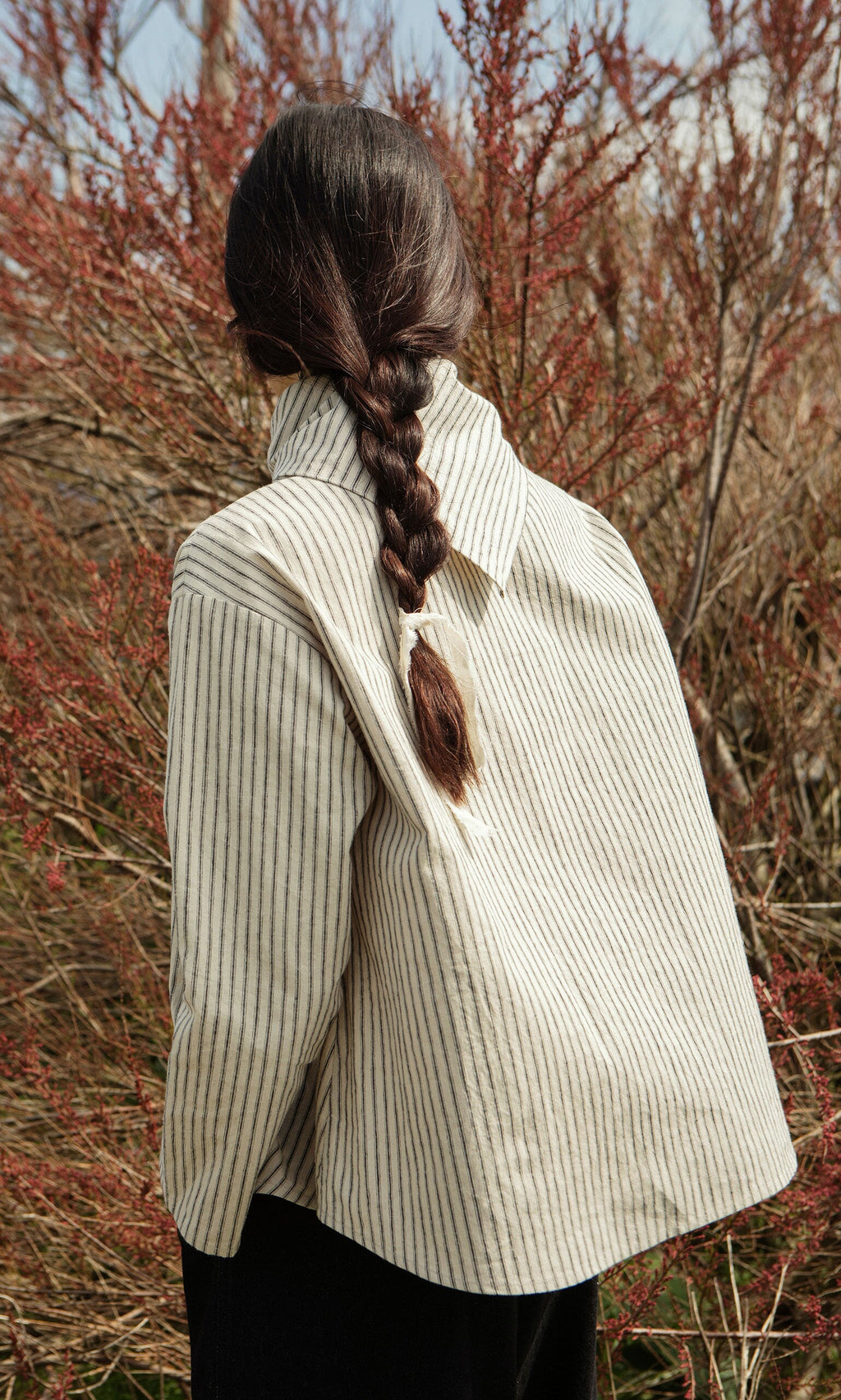 Plümo- Person with long brown braid stands outdoors in a Sail Cloth Top, back turned, before bare reddish-brown bushes.