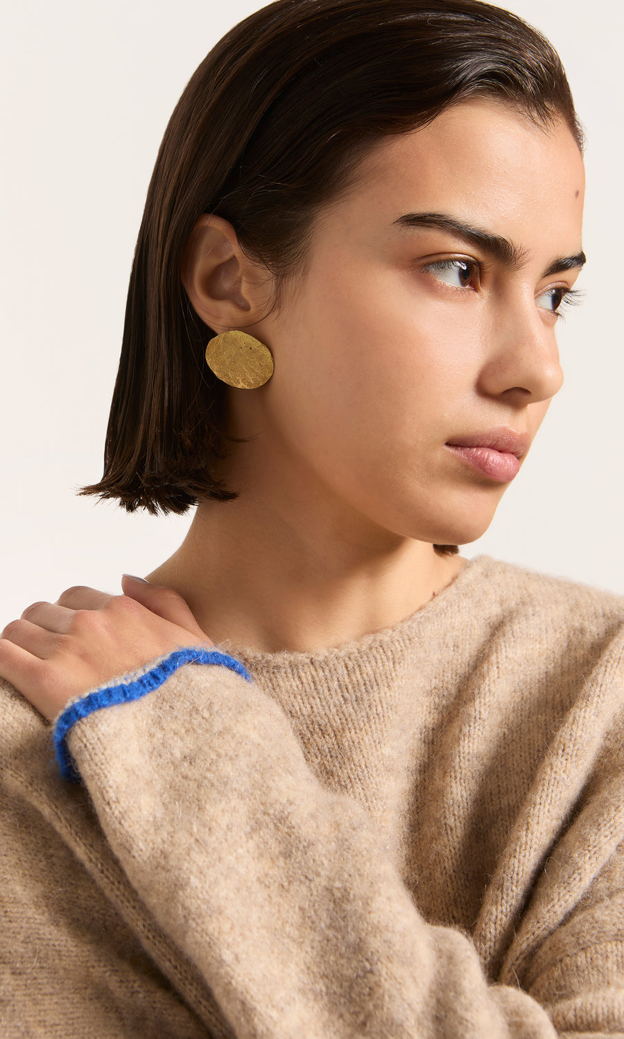 Plümo  Woman in a beige sweater with blue trim wears handmade Cohen Earrings—large gold ovals—looking to the side.