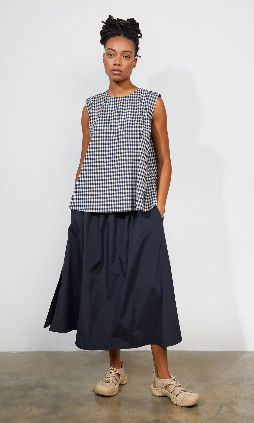 Plümo- Woman wearing the Serena Top - Gingham, a navy midi skirt, and beige sandals, standing against a plain wall.