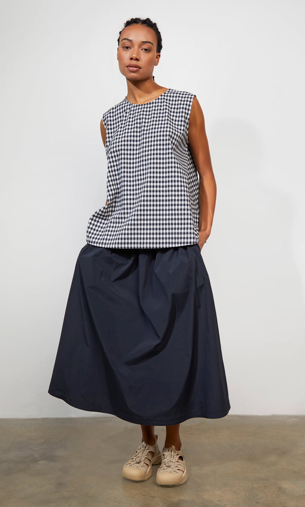Plümo- Woman in Serena Top - Gingham, navy maxi skirt, and beige sneakers, standing indoors.