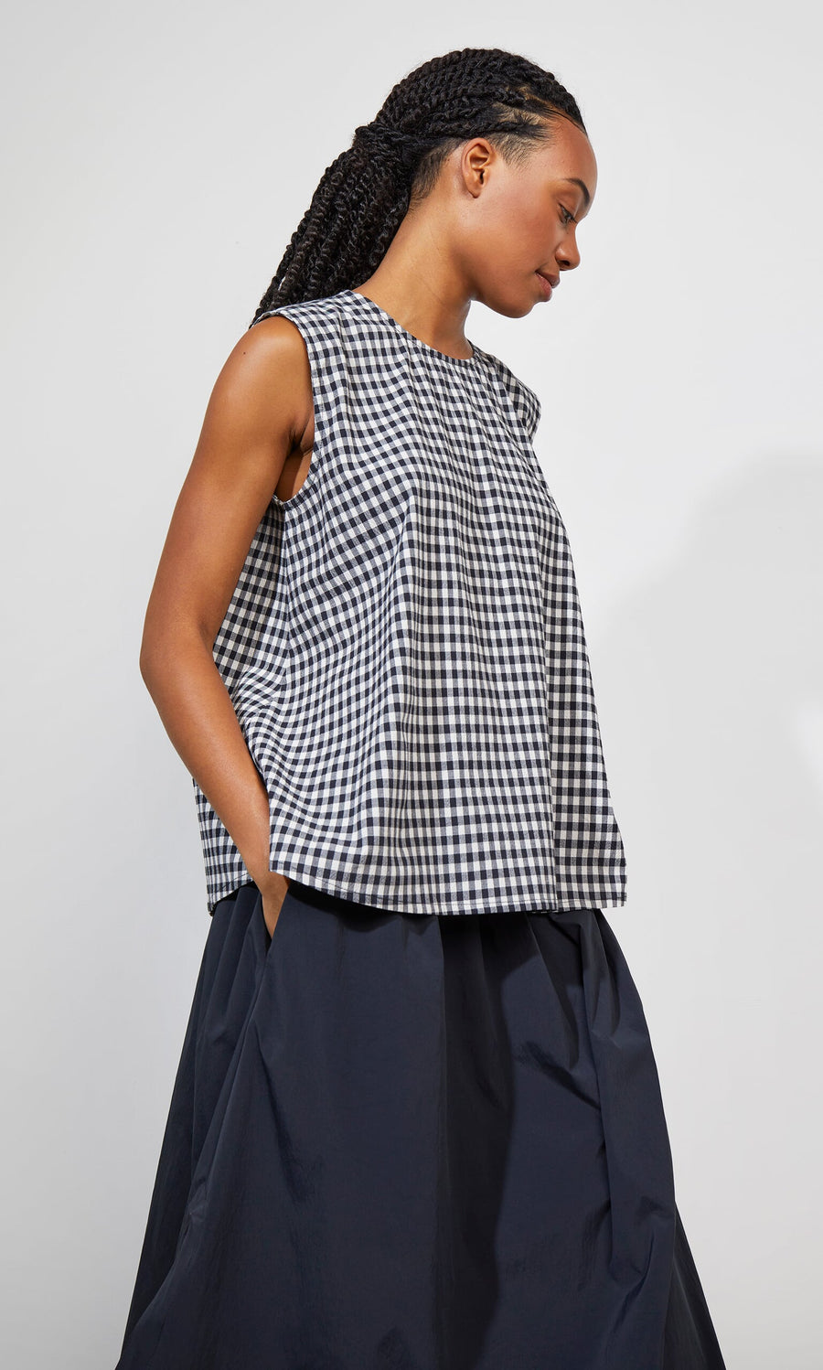 Plümo- Woman in the Serena Top - Gingham, a sleeveless seersucker trapeze style, and a dark skirt, standing sideways.