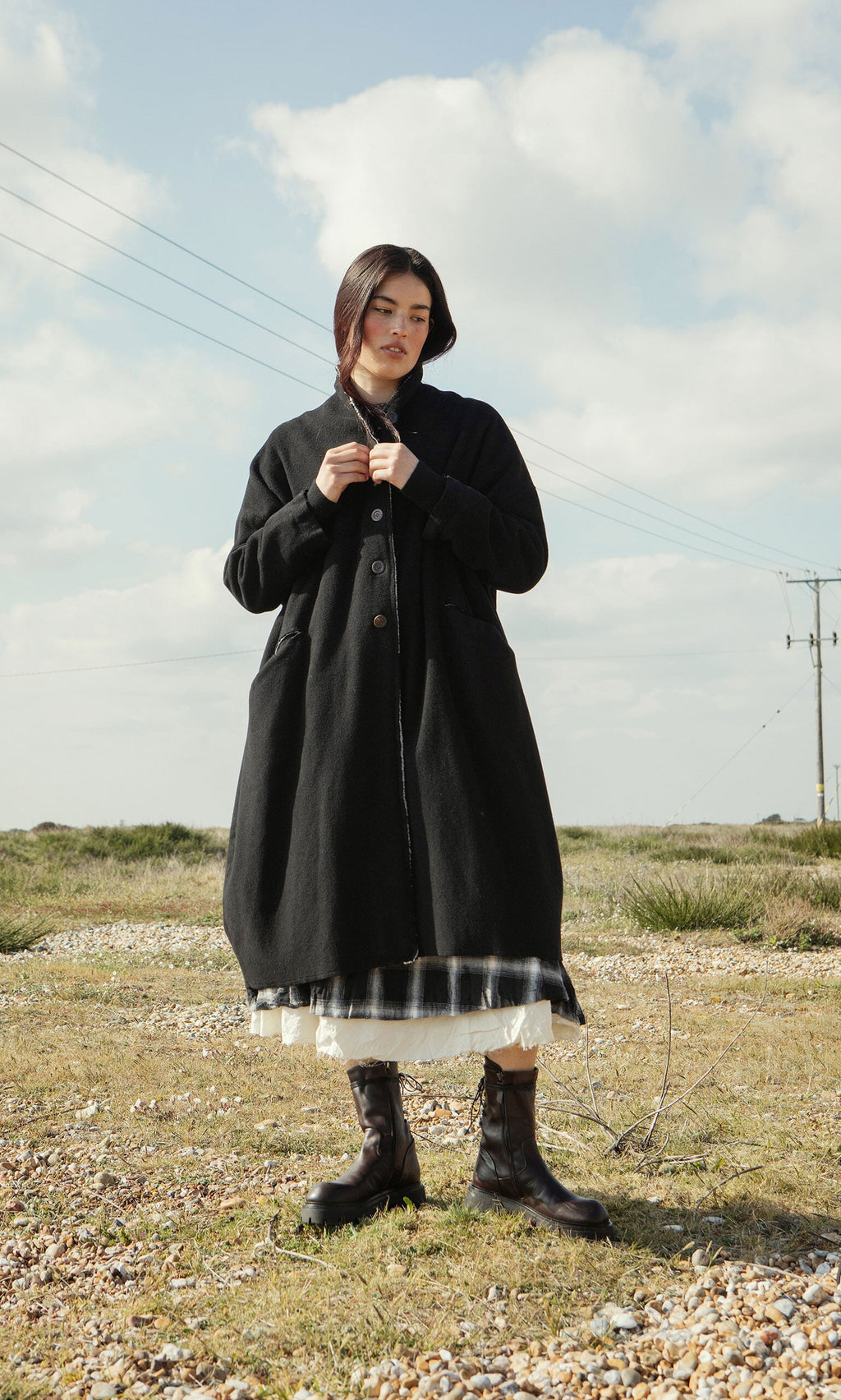 Plümo- A woman in the Sigrun Coat and boots stands on grass and pebbles beneath a cloudy sky.