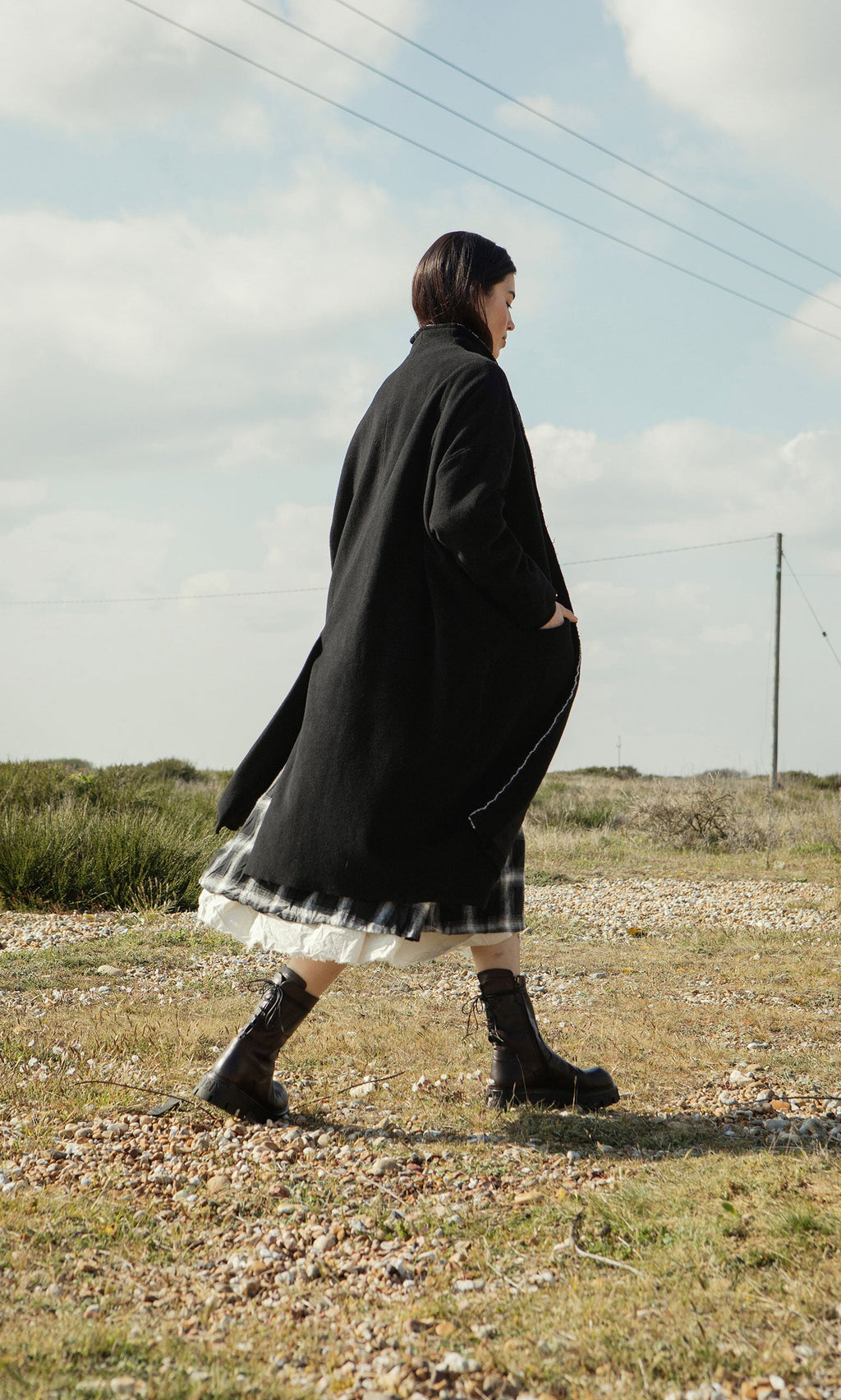 Plümo- Wearing the Sigrun Coat, a person in a layered skirt and boots walks outdoors on a grassy, rocky path under cloudy skies.