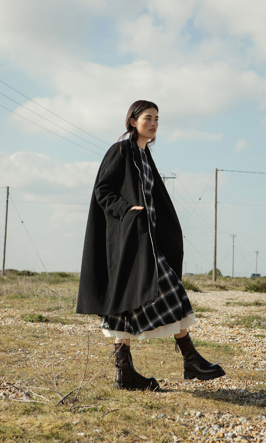 Plümo- A woman in the Sigrun Coat and boots walks outdoors on a grassy, rocky field under a partly cloudy sky.