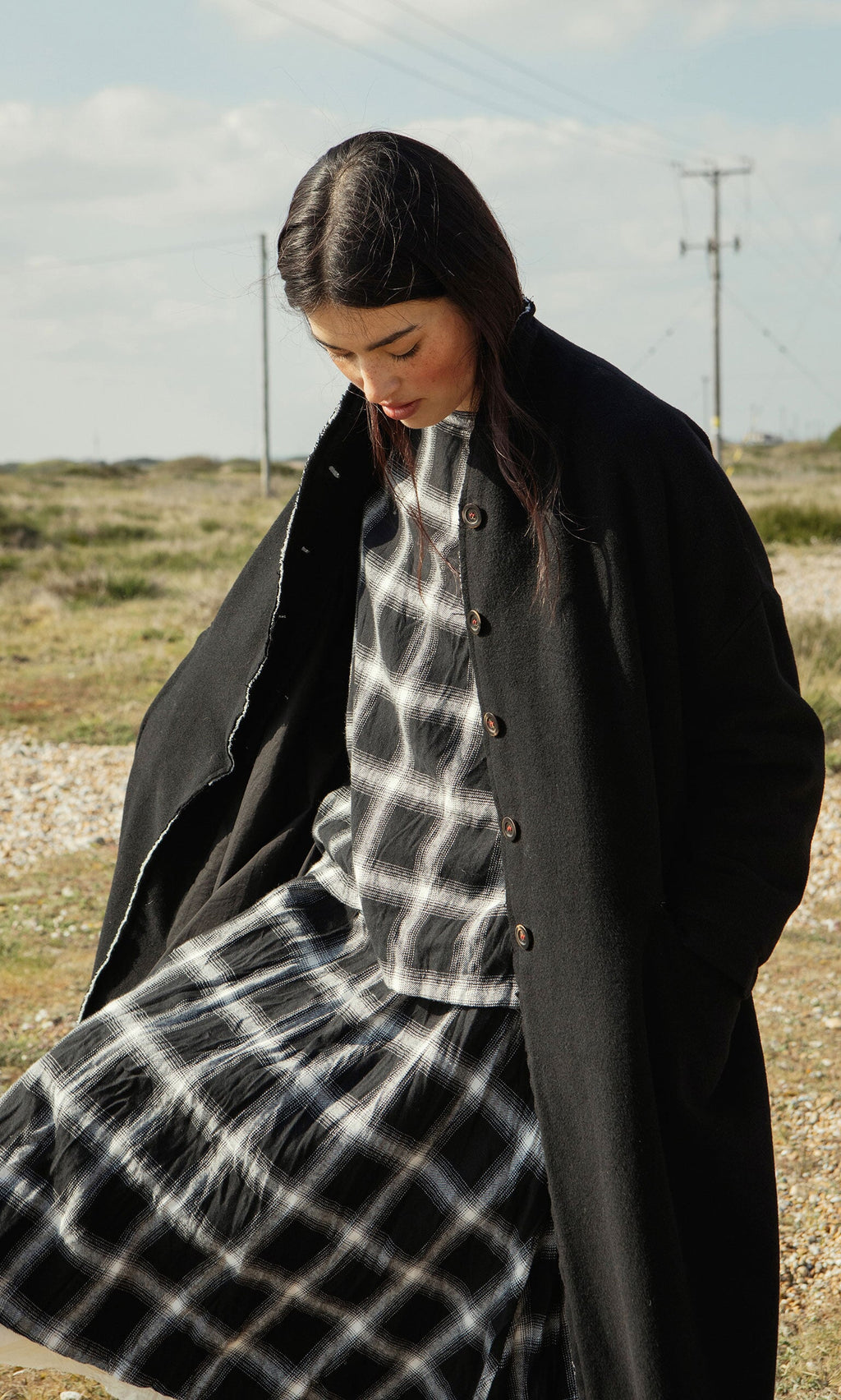 Plümo- Woman outdoors in the Sigrun Coat, a black wool-cashmere design, with hands in pockets over a plaid set.