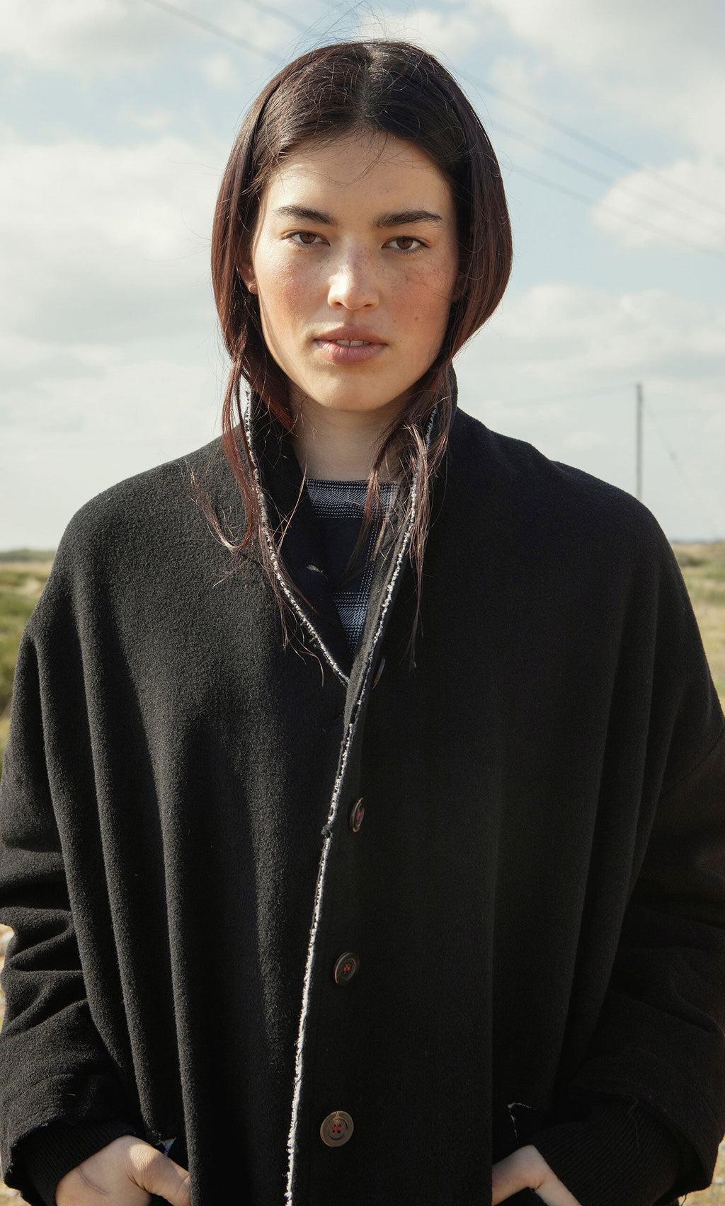 Plümo- A person with long dark hair wears the Sigrun Coat and stands outdoors under a cloudy sky, looking at the camera.