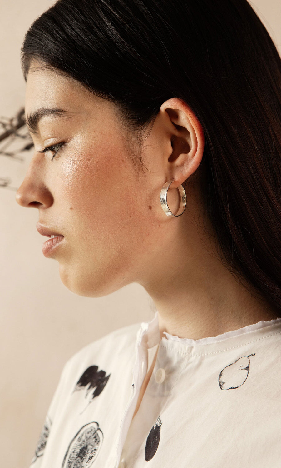 Plümo- Woman with long dark hair wears Silver Pebble Hoops and a white shirt with black patterns, shown in side profile.