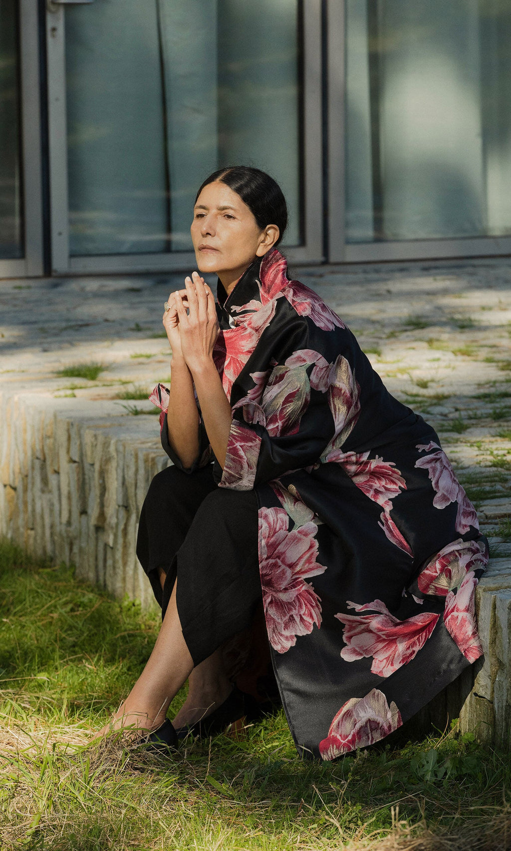 Plümo- A woman in a Rozella Coat sits outdoors on a stone ledge, hands clasped, looking thoughtful.