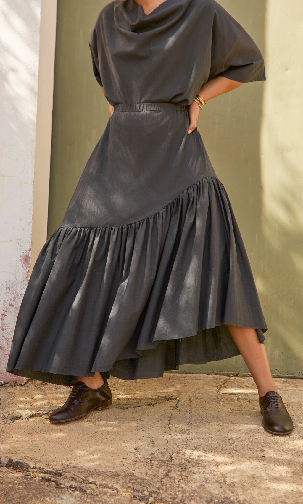 Plümo- A person in a dark Skegg Skirt from PLÜMO Studio stands hands on hips on a sunlit outdoor path.
